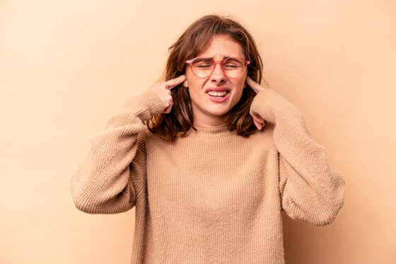young caucasian woman isolated beige background covering ears with fingers stressed desperate by loudly ambient 560x373jpg